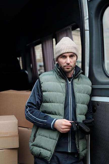 A man wearing a light grey knit beanie, a green padded vest, and a dark zip-up jacket with striped sleeves stands inside a van during a home relocation process. Behind him, cardboard boxes are stacked, some wrapped in protective plastic or fabric for transport. The van's interior is visible, with windows along the side, and part of the vehicle's door frame. The man appears to be holding a strap or securing item, likely preparing for loading or unloading furniture or boxes. This scene relates to the logistics of packing and moving, typical of house removals services such as those offered by Man and Van Southfields, focusing on efficient furniture transport and the loading process in a home relocation context.