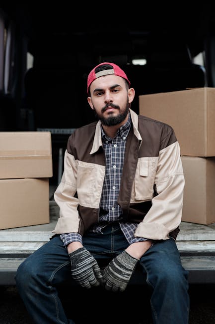 A young man with a beard is seated on the ground at the back of a moving van, surrounded by brown cardboard boxes of various sizes. He is wearing a red cap backwards, a checkered shirt underneath a beige and brown jacket, and striped gloves. The interior of the van is visible behind him, with additional boxes and dark upholstery. The scene depicts the loading process involved in home relocation or furniture transport, typical of professional removals services such as those offered by Man and Van Southfields. The natural lighting highlights the details of the packing materials and the man's focused expression while preparing for moving logistics.