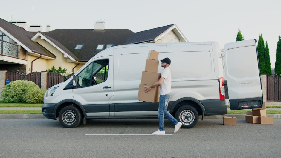 A man wearing a light grey knit beanie, a green padded vest, and a dark zip-up jacket with striped sleeves stands inside a van during a home relocation process. Behind him, cardboard boxes are stacked, some wrapped in protective plastic or fabric for transport. The van's interior is visible, with windows along the side, and part of the vehicle's door frame. The man appears to be holding a strap or securing item, likely preparing for loading or unloading furniture or boxes. This scene relates to the logistics of packing and moving, typical of house removals services such as those offered by Man and Van Southfields, focusing on efficient furniture transport and the loading process in a home relocation context.