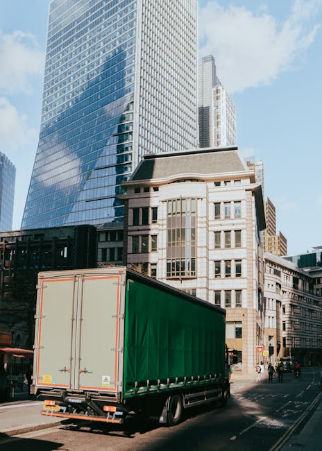 A large green and cream box truck is parked on a city street in Southfields, with tall modern office buildings made of glass and concrete rising behind it. The truck is positioned near the curb, with its rear doors closed, ready for loading or unloading. The setting is during the daytime with a clear sky, and there are no visible moving activity happening at this moment. The scene captures urban surroundings typical of a home relocation or furniture transport process, reflecting the logistical aspect of house removals in Southfields. Man and Van Southfields offers professional moving services, including vehicle transport for house moves, as evidenced by the truck parked ready for a potential house relocation or move logistics nearby.