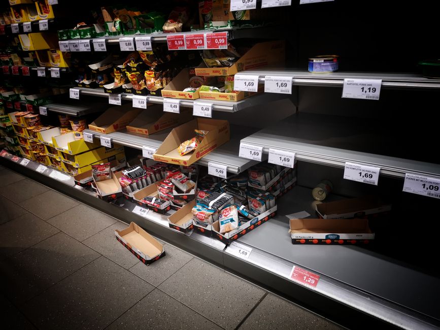 Empty supermarket shelf in a grocery store aisle with partially stocked cardboard boxes and loose snack packets on the lower tier, while the upper shelves display various packaged food items and snack