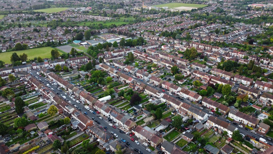 An aerial view of a residential neighbourhood in Southfields, showing multiple rows of terraced houses with pitched roofs, small front gardens, and parked cars lining the streets. The image captures a broad perspective of the area, including green open spaces and trees interspersed among the houses. In the foreground, a street is visible with closely parked vehicles, and the houses feature typical British terraced architecture with brick facades and various roof materials. The surrounding environment includes well-maintained gardens, pathways, and some larger communal green areas suitable for home relocation planning or furniture transport in SW18. This detailed overview of a typical suburban street provides insight into the logistical considerations involved in house removals, supported by the professional services offered by Man and Van Southfields.