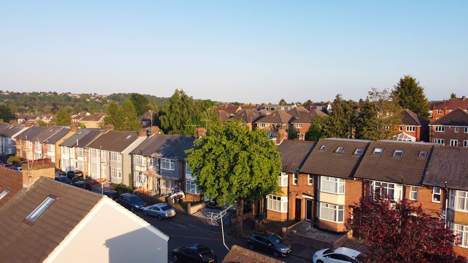 An aerial view of a residential neighbourhood in Southfields, featuring a row of terraced houses with pitched roofs, some with skylights. In the foreground, a large leafy green tree partially shades the street below, where several parked cars are lined up along the road and pavement. The street appears calm and clean, with sidewalks on each side. The sky above is clear and blue, indicating good weather, and the houses are set among other similar homes with trees and greenery in the background. This scene visually represents a typical suburban area suitable for house removals and relocation services, with the focus on typical residential properties and moving logistics.