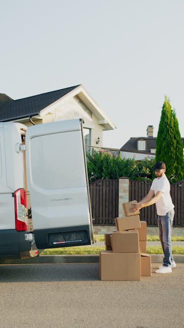 A man with a beard, wearing a white t-shirt, jeans, and white sneakers, is carrying three stacked cardboard boxes while unloading a white cargo van parked on a suburban street. The van's side door is