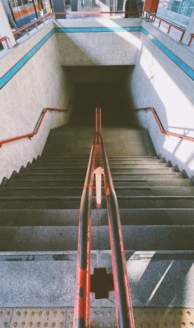 The image shows an indoor staircase with concrete steps and orange metal handrails on both sides, leading down from a well-lit corridor to an underground passage. The walls are made of grey textured material, with a blue border near the top, and large windows at the top allowing natural light to illuminate the area. Visible in the background is an entry vestibule with a glass door and railing, indicating the stairway is part of a public transportation station or building. This setting captures the typical environment for passenger movement or access during home relocation or transport processes, aligning with house removals and furniture transport considerations. Man and Van Southfields often utilizes such stairways when navigating tight street access or underground entry points during residential moving services, ensuring careful handling of furniture and boxes in alignment with relocation logistics.