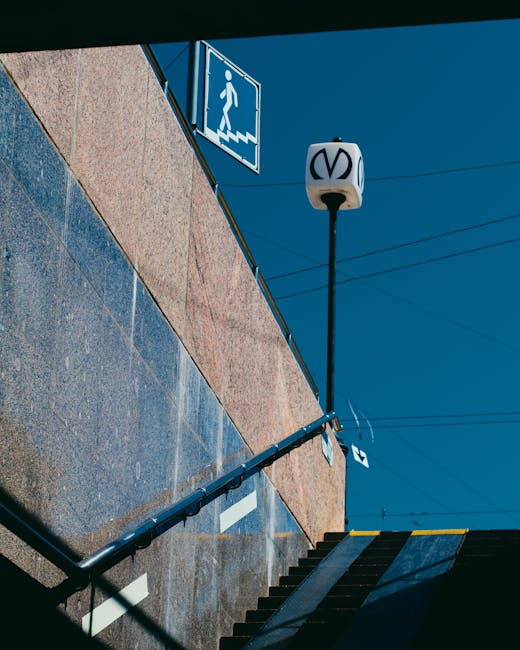 A set of outdoor concrete stairs leading down from a street level entrance, with a black metal handrail on the right side. Adjacent to the stairs is a brown brick wall, topped by a beige-colored wall. Above the stairs, mounted on the wall, is a blue and white sign indicating a pedestrian staircase with an icon of a walking person and stairs. A street lamp with a white, rounded fixture featuring a black logo is attached to a tall black pole above the stairs, extending upward into a clear blue sky. The scene suggests an urban environment near Southfields station, with overhead electrical wires visible. This setting could be associated with a home relocation or furniture transport process handled by Man and Van Southfields, especially relevant to moving in or out through tight street access, as indicated in the page about Southfields station removals tips.