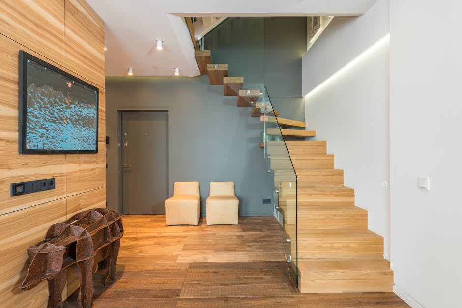 The image depicts a modern interior entrance of a residential property with a staircase leading to the upper floor. The staircase is made of light-colored wood with glass panel balustrades on one side and wooden steps attached to a gray wall. Two beige upholstered armchairs are positioned against the gray wall at the bottom of the stairs, near a closed gray door. On the left side, there is a wooden accent wall with a mounted flat-screen television displaying an abstract blue and black pattern, and an image of two carved wooden animal figures is placed on the floor beneath it. Illuminated ceiling spotlights shine down, providing ample lighting for the space. The environment appears clean, organized, and suitable for house removals or furniture transport, with the minimalistic design emphasizing the ease of moving large furniture items through the stairway during a home relocation process. Man and Van Southfields may utilise this layout for efficient packing and loading within this modern apartment setting.