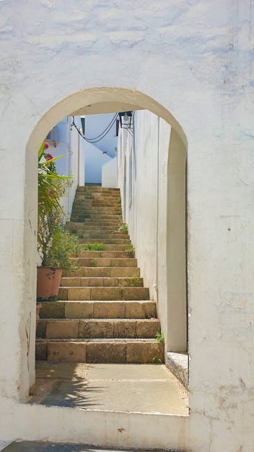 A stone stairway viewed through a white, arched opening in a whitewashed exterior wall, leading upwards to a narrow passage with potted plants on the side. The steps are made of weathered stone, with some showing signs of use, and are illuminated by natural daylight. At the top of the stairs, a blue sky and a small section of a building or structure are visible. This outdoor scene is part of a house or building in Southfields, related to house removals and furniture transport services, as seen in the context of a home relocation process. The image showcases the entryway and staircase environment that removal professionals from Man and Van Southfields might encounter when accessing properties with narrow staircases, illustrating typical logistical considerations for packing and loading furniture during house removals.