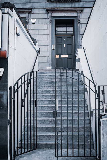 The image depicts a modern interior entrance of a residential property with a staircase leading to the upper floor. The staircase is made of light-colored wood with glass panel balustrades on one side and wooden steps attached to a gray wall. Two beige upholstered armchairs are positioned against the gray wall at the bottom of the stairs, near a closed gray door. On the left side, there is a wooden accent wall with a mounted flat-screen television displaying an abstract blue and black pattern, and an image of two carved wooden animal figures is placed on the floor beneath it. Illuminated ceiling spotlights shine down, providing ample lighting for the space. The environment appears clean, organized, and suitable for house removals or furniture transport, with the minimalistic design emphasizing the ease of moving large furniture items through the stairway during a home relocation process. Man and Van Southfields may utilise this layout for efficient packing and loading within this modern apartment setting.