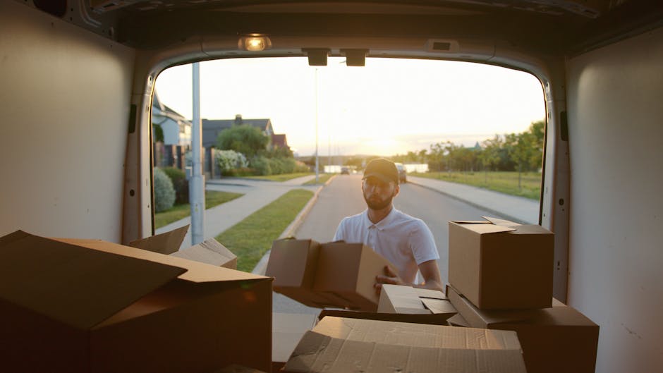A male and female mover are engaged in a home relocation process outside a residential building with brick exterior walls and large windows. The male mover, wearing a maroon t-shirt, is smiling and le