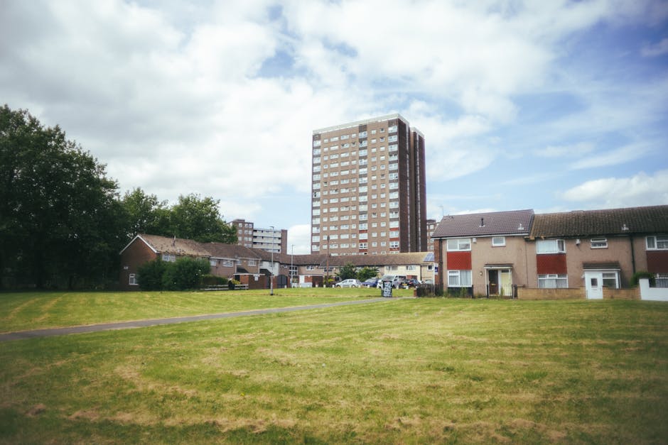 A wide view of a residential area with a large grassy field in the foreground and a row of single-story houses with brown tiled roofs and light-colored walls behind it. In the background, a tall multi-storey apartment building with numerous windows and a brick facade rises above the smaller houses. The sky is partly cloudy with patches of blue, and some trees are visible on the left side. Several vehicles are parked on the street near the houses. The scene appears to be taken during the daytime, and the environment suggests a suburban setting suitable for home relocation or furniture transport, as coordinated by Man and Van Southfields for removals services.
