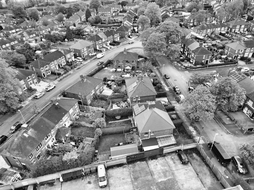 An aerial black and white photograph showing a suburban residential area with multiple detached and semi-detached houses, each with small gardens and yards. Several houses have visible roofing materials such as tiles and metal sheets. A narrow street runs through the neighbourhood, with parked cars lining both sides and a few moving vehicles. Trees with full foliage are dispersed throughout the area, providing shade and greenery. In the foreground, a flat roof area with a few scattered items and a small fenced garden can be seen. The surroundings feature other streets and clusters of houses extending into the distance, with visible boundary fences, driveways, and small communal pathways. This setting depicts a typical UK residential neighbourhood suitable for home relocation or furniture transport, where a moving company like Man and Van Southfields might assist with loading and transport logistics amidst everyday urban living environments.