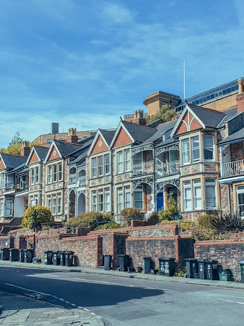 A row of Victorian-style terraced houses with decorative ironwork balconies and bay windows, situated behind a brick wall and pavement. Several black wheelie bins are lined up along the street in front of the houses. The sky is clear with minimal clouds, and the scene is well-lit, suggesting daytime. This setting indicates an urban residential area where house removals and furniture transport by Man and Van Southfields may take place, especially during home relocation or packing and moving processes within Southfields, aligning with the context of Wandsworth Council removal permits.