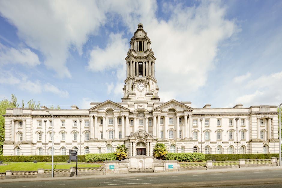 A historic stone building with a central clock tower, featuring ornate architectural details and a symmetrical façade with multiple windows. The building is situated on a street with a paved road in the foreground, and landscaped greenery including bushes and small trees in front. The clock tower has a prominent, pagoda-like spire with open arches and decorative elements. In the context of house removals or home relocation, this image may represent a notable landmark or council building relevant to Wandsworth Council removal permits for Southfields. The sky is partly cloudy with patches of blue, and the scene is well-lit with natural daylight, emphasizing the building's classical architecture and decorative stonework. Occasionally, [COMPANY_NAME] undertakes furniture transport and packing processes near such structures during residential moving services, ensuring efficient loading and safeguarding of household goods for property transitions.