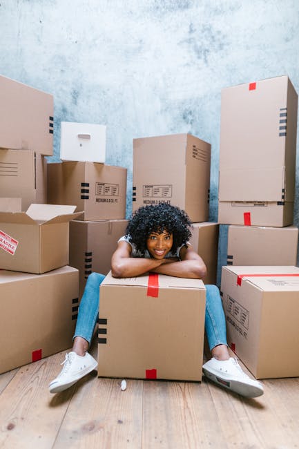 A young woman with curly black hair sits on a wooden floor inside a room surrounded by numerous packed cardboard boxes, some open with flaps folded back. The boxes vary in size and are sealed with red or clear packing tape, with some displaying shipping labels and barcodes indicating they are prepared for a house relocation. The woman is smiling and resting her chin on a medium-sized box positioned directly in front of her, with her arms draped over it. The room has a textured light blue wall and appears to be part of a residential property, with no furniture visible except for the boxes. The scene captures the loading process typical of furniture transport and packing during a same-day removal service, with the boxes and the woman representing a house move organized for efficiency and quick relocation, as provided by companies like Man and Van Southfields. Bright lighting highlights the details of the packed items, emphasizing the logistics involved in homestorage and furniture transport.