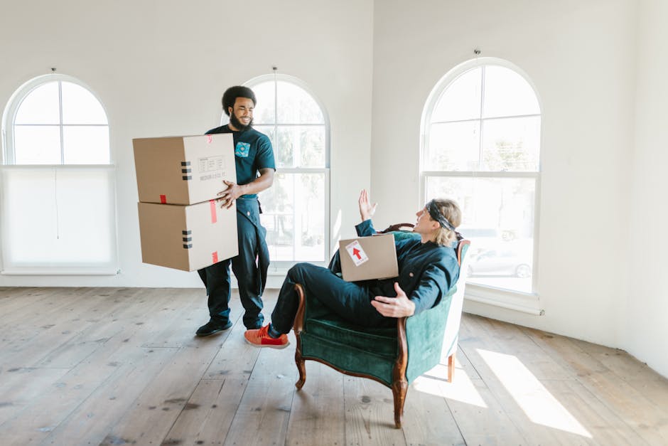 A man with a dark beard, wearing a navy blue uniform, is inside a bright, spacious room with large arched windows that let in natural light. He is holding two cardboard boxes, one stacked on top of the other, secured with red and black packing tape, and appears to be preparing to load or transport them. A woman with glasses, light brown hair, and dressed in dark clothing is seated in an antique green armchair with wooden legs. She is engaging with the man, raising one hand and holding a medium-sized cardboard box with a white arrow label on it across her lap. The room features wooden flooring and minimal interior decor. Behind them, the windows reveal a glimpse of an outdoor suburban street. The scene captures a moment during the home relocation process, involving packing, lifting, and moving household items, consistent with services offered by Man and Van Southfields for efficient furniture transport and same-day removals.
