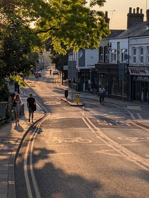 A quiet street scene during late afternoon with sunlight casting long shadows on the road. The street features double yellow lines along the curb, with a pedestrian crossing and a central traffic island topped with a yellow bollard. On the left side, two people are walking along the pavement, while on the right, a cyclist rides past a shopfront with a dark exterior and large windows. A large tree with green foliage extends its branches across the scene, partially shading the road. In the background, there are parked cars, additional buildings, and a clear sky. The scene illustrates a typical urban environment suitable for residential or commercial relocation activities, as part of home relocation or furniture transport arrangements. Man and Van Southfields might be involved in such moving logistics, supported subtly by the calm and organized street setting.